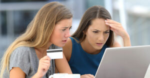 Two young women reviewing a charge on a laptop, holding a credit card with concerned expressions.
