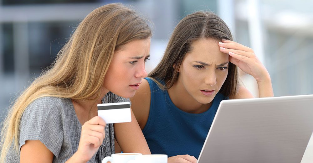 Two young women reviewing a charge on a laptop, holding a credit card with concerned expressions.